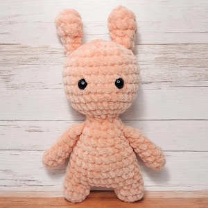 May include: A peach-colored, handmade crochet bunny with black safety eyes. The plush toy has a rounded head, long ears, and small arms and legs. The bunny is made of a soft, textured yarn and is displayed against a light wood background.