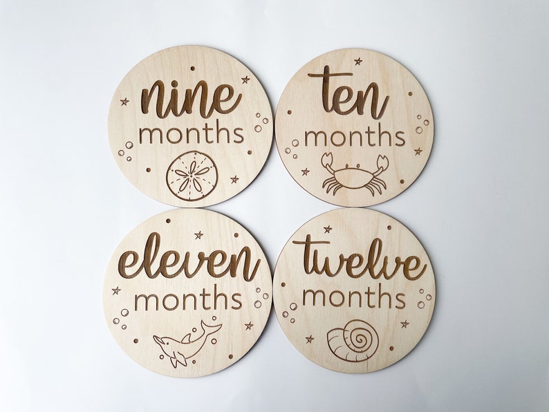 May include: Four circular wooden milestone plaques with engraved text and marine illustrations. The plaques display "nine months", "ten months", "eleven months", and "twelve months". Each features a different sea element: a sand dollar, crab, dolphin, and seashell.