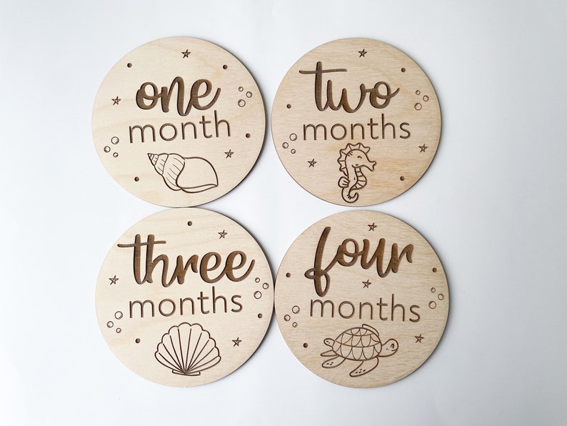 May include: Four wooden baby milestone discs with engraved text and sea-themed illustrations. The discs are labelled "one month," "two months," "three months," and "four months." Each disc features a different sea creature: a seashell, seahorse, seashell, and turtle.