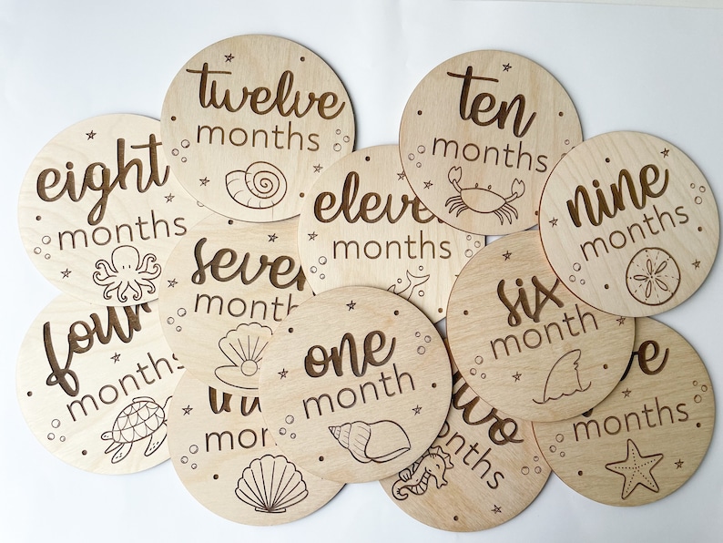 May include: A set of wooden monthly milestone discs. Each disc is round and engraved with a month number and a sea-themed illustration, like a seashell or starfish. The discs are made of light-coloured wood.