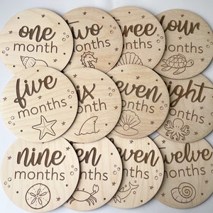 May include: A collection of twelve wooden milestone discs, each marked with a month number and the word "months". The discs are round and feature sea-themed illustrations, such as a starfish and a seashell, in a dark brown color.
