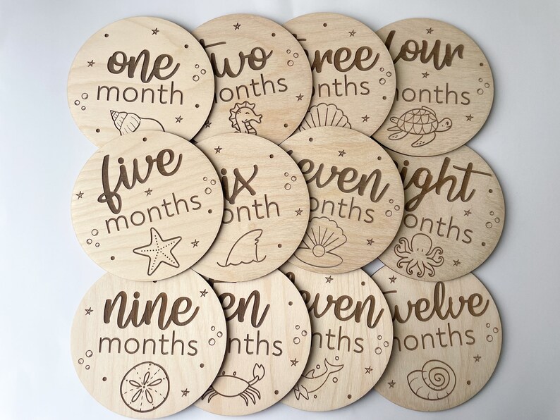 May include: A collection of twelve wooden milestone discs, each marked with a month number and the word "months". The discs are round and feature sea-themed illustrations, such as a starfish and a seashell, in a dark brown color.