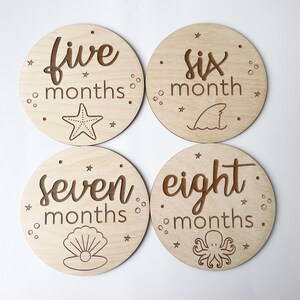 May include: Four circular wooden milestone discs with engraved text and marine illustrations. The discs are marked "five months," "six month," "seven months," and "eight months." Each disc features a different sea creature or element.