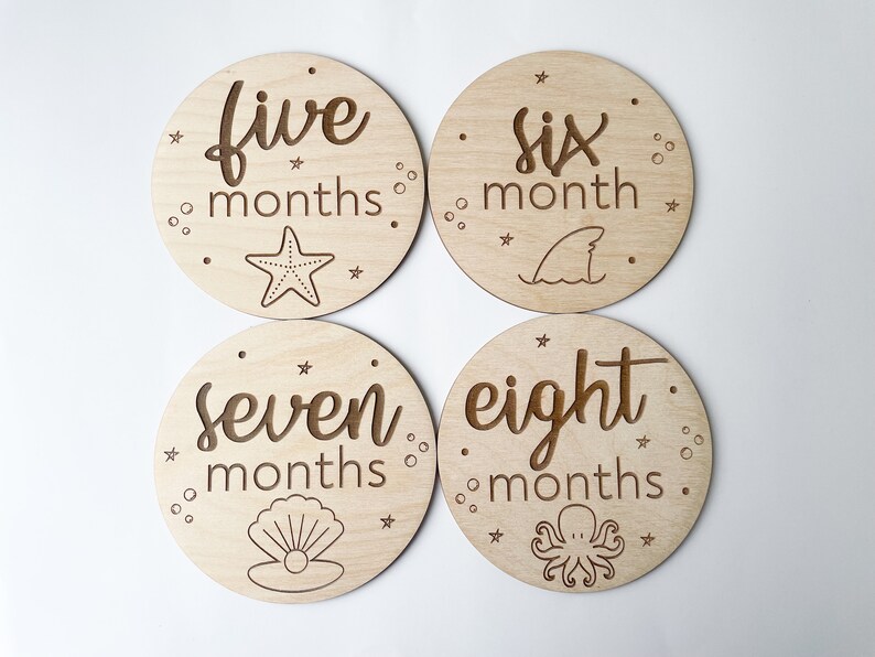 May include: Four circular wooden milestone discs with engraved text and marine illustrations. The discs are marked "five months," "six month," "seven months," and "eight months." Each disc features a different sea creature or element.