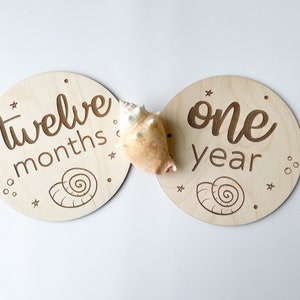 May include: Two circular wooden milestone plaques with engraved text and seashell designs. One says "twelve months," the other "one year." A seashell sits between them. Ideal for marking a baby's first year.