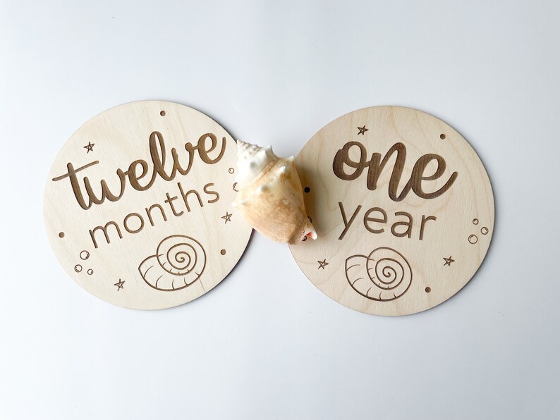 May include: Two circular wooden milestone plaques with engraved text and seashell designs. One says "twelve months," the other "one year." A seashell sits between them. Ideal for marking a baby's first year.