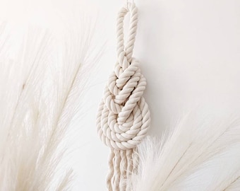 May include: A hanging wall ornament crafted from thick, off-white twisted cotton rope. The rope is intricately tied into a complex, multi-layered decorative knot at the center, featuring a loop at the top and several loose, frayed rope ends at the bottom.