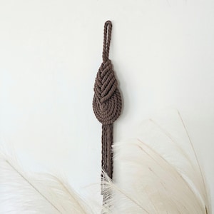 May include: A brown macrame wall hanging with a tassel. The hanging is made of thick rope and has a knotted design.