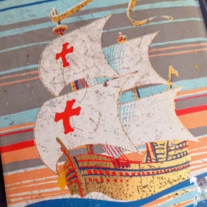 Può includere: Scatola rettangolare a tema nautico. Il coperchio presenta un'illustrazione dettagliata di una nave a vela con vele bianche, croci rosse e scafo dorato, su uno sfondo di strisce blu e arancioni e onde blu.