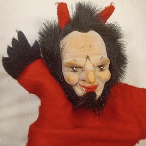 May include: A vintage devil hand puppet with a red felt body and face. The puppet has a painted face with a prominent nose, red lips, and black beard. It features red horns and black fur around the face.