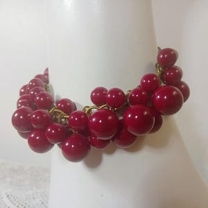 Può includere: Un braccialetto vintage con grappoli di grandi perle rotonde bordeaux. Le perle sono infilate con maglie di catena dorate. Il braccialetto è esposto su una forma bianca, mettendo in risalto il suo design e colore.
