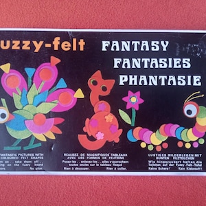 Op de afbeelding: Een rechthoekige zwarte doos voor een fuzzy-felt set met de tekst fuzzy-felt FANTASY FANTASIES PHANTASIE. De doos toont kleurrijke viltvormen gerangschikt als een vogel, een kat, een bloem en een rups op een zwarte achtergrond.
