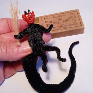 May include: A small devil figurine crafted from black pipe cleaners with a red painted head, black horns, and a long tail. The figure holds a miniature broom made of light-coloured straw. Beside it sits a rectangular matchbox labelled MANUFACTURES DE L'ETAT.