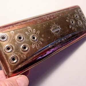 May include: A rectangular brass harmonica featuring a metallic finish with engraved floral patterns and circular sound holes. The centre of the instrument displays a small sunburst emblem with the text "M. HOHNER" clearly visible in the middle.