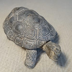 Peut inclure: Figurine décorative de tortue grise avec une carapace texturée et des pattes détaillées. La figurine a un aspect vieilli, avec un mélange de gris et de tons blanc cassé. La tortue est posée sur une surface de couleur claire.