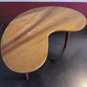May include: A mid-century modern kidney-shaped coffee table. The table is made of wood with a light brown finish and darker wood grain patterns. It has three short, tapered legs. The table measures approximately 76 cm long.