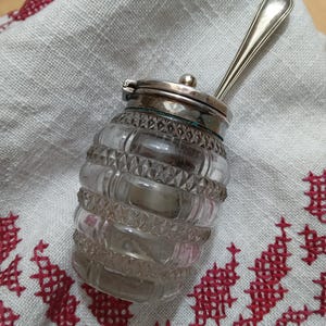 Puede incluir: Un tarro de cristal transparente con tapa y cuchara plateadas. El tarro tiene un diseño decorativo y texturizado. La cuchara descansa sobre la tapa. El tarro está sobre un paño blanco con bordado de punto de cruz rojo.