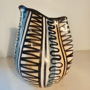 Puede incluir: Una jarra de cerámica con cuerpo redondeado y borde asimétrico. El exterior está decorado con paneles verticales de líneas negras en zigzag, franjas de color azul claro y bandas de color amarillo pálido sobre un fondo blanco.