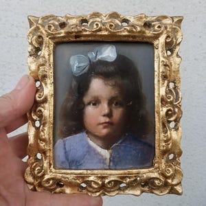 May include: A rectangular portrait painting of a child with dark hair and a light blue bow, wearing a blue garment. The artwork is encased in an ornate, textured gold-coloured frame with intricate scrollwork details along the edges.