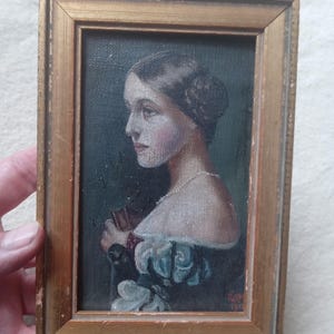 May include: An oil painting in a distressed gold-toned wooden frame depicting a profile portrait of a person with dark hair styled in a bun, wearing an off-the-shoulder garment with blue and white accents, signed and dated 1911 in the bottom corner.