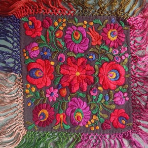 Könnte beinhalten: Ein quadratisches, dunkelbraunes Stoffstück mit dichter, bunter Blumenstickerei in Rot-, Rosa-, Orange-, Blau- und Grüntönen. Die Kanten sind mit langen, mehrfarbigen Seidenfransen in Rot, Gold, Blau, Rosa und Grün versehen.
