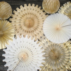Könnte beinhalten: Eine Sammlung von goldenen und weißen Papierfächern, einige mit gewellten Rändern und einige mit ausgeschnittenen Designs. Die Fächer sind auf einer Holzoberfläche angeordnet.