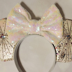 May include: Ivory-colored headband with sequined ears and a large bow. The ears feature a radial design with silver and colorful bead accents. The bow and ears are covered in iridescent sequins. The headband is white.