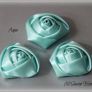 Könnte beinhalten: Drei aquablaue Satinbandrosen. Die Rosen sind in einer Reihe auf einer grauen Oberfläche angeordnet. Der Text "Aqua" ist in Schwarz auf der linken Seite des Bildes geschrieben. Der Text "All Season Bloom Supply" ist in Schwarz auf der rechten Seite des Bildes geschrieben.