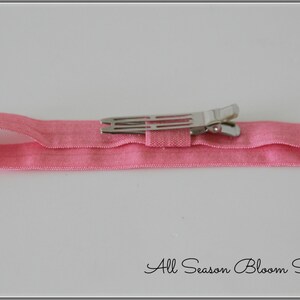 Pu&ograve; includere: Un cerchietto elastico rosa con una clip a coccodrilo argentata attaccata. Il cerchietto &egrave; piegato a met&agrave; e la clip &egrave; chiusa.