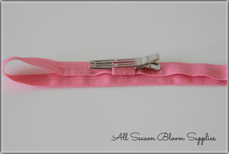 Pu&ograve; includere: Un cerchietto elastico rosa con una clip a coccodrilo argentata attaccata. Il cerchietto &egrave; piegato a met&agrave; e la clip &egrave; chiusa.