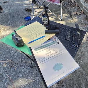 Op de afbeelding: Een zwarte klaptafel toont een veldgids, portofoons, een verrekijker, een mes en een kompas. De veldgids heeft de titel "The Cryptid Field Companion" en bevat de tekst "A Practical Awareness Guide for Campers & Explorers."