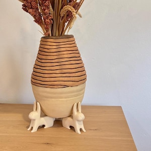 May include: A ceramic vase with a light brown, horizontally striped upper section and a cream-colored base. The vase is supported by three small, white bunny figurines. The vase holds dried flowers and grasses.