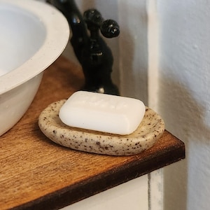 May include: A miniature bathroom scene featuring a white bar of soap resting on a speckled soap dish. The dish is set on a wooden surface, with a black faucet and a white sink visible in the background. The soap dish has a speckled pattern.