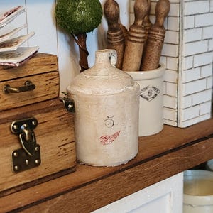 May include: A miniature beige stoneware jug featuring a prominent number 5 and a small red wing logo on the front. The jug has a cylindrical body, a short neck with a handle, and a textured finish, displayed alongside wooden boxes and a crock.