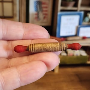 May include: A miniature rolling pin with a light brown wooden barrel and red handles. The small kitchen tool is designed for a dollhouse or other miniature setting. The rolling pin is held in a person's hand.