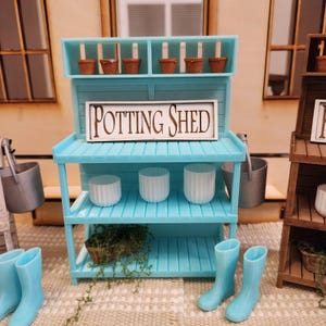 May include: A miniature potting shed in light blue with the words "Potting Shed" on a sign. The shed has shelves with small pots, white planters, and a basket of greenery. Blue rain boots are in the foreground.