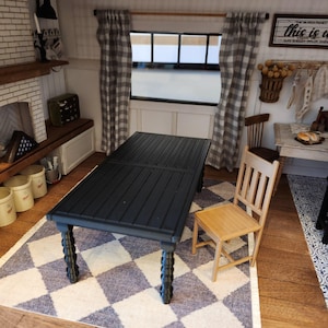 May include: A miniature black dining table with a slatted top and ornate legs sits on a blue and white checkered rug. A light wood chair is positioned next to the table. The scene is set in a dollhouse kitchen.