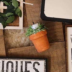 May include: A small, orange terracotta pot with a white candle and green moss. The pot is tilted, and the candle has a black wick. The pot is on a wooden surface, with other decorative elements in the background.
