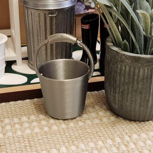 May include: A small, silver-colored bucket with a handle. The bucket is cylindrical with a slightly flared top. It is sitting on a beige and white patterned surface. Other miniature items are in the background.