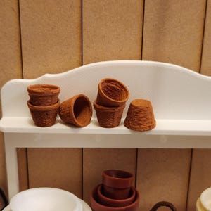May include: A collection of small, brown miniature flower pots, arranged on a white shelf. The pots have a woven basket design. The background features a light brown wooden paneling.