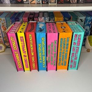 May include: A series of brightly coloured, handmade book-shaped boxes with amusing phrases printed on the spines. The boxes are arranged in a row, each with a different colour and text, including phrases like "MONGO IS APPALLED!" and "IT'S LIKE HIRING AN INCUBUS".