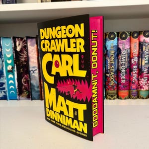 Op de afbeelding: Een hardcover boek getiteld "Dungeon Crawler Carl" van Matt Dinniman. De omslag is zwart met gele tekst en een roze rug. Het boek wordt weergegeven op een witte plank met andere boeken op de achtergrond.