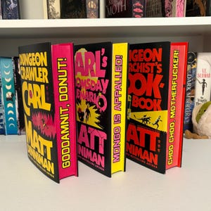 May include: A selection of hardback books with striking, vibrant covers. Titles visible are "Dungeon Crawler Carl", "Goodamnit, Donut!", "Arl's Doomsday Enario", "Mongo is Appalled!", "Dungeon Archist's Cookbook", and "Choo Choo, Motherfucker!"