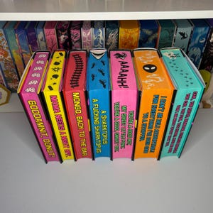 May include: A collection of colorful, decorative book-shaped boxes with various phrases and graphics. The boxes are arranged in a row, with text like "Gooddamnit, Donut!" and "A Sharktopus" visible. The boxes are on a white surface.