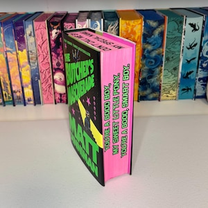 May include: A hardcover book titled "The Butcher's Masquerade" by Matt Goldman. The book has a black cover with neon green and pink accents. The spine is bright pink with text. Other books are visible on a shelf in the background.