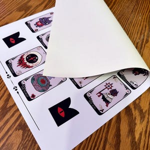 May include: A set of tarot cards featuring illustrations of various characters and symbols. The cards are printed on white paper with black borders. The cards are laid out on a wooden surface.