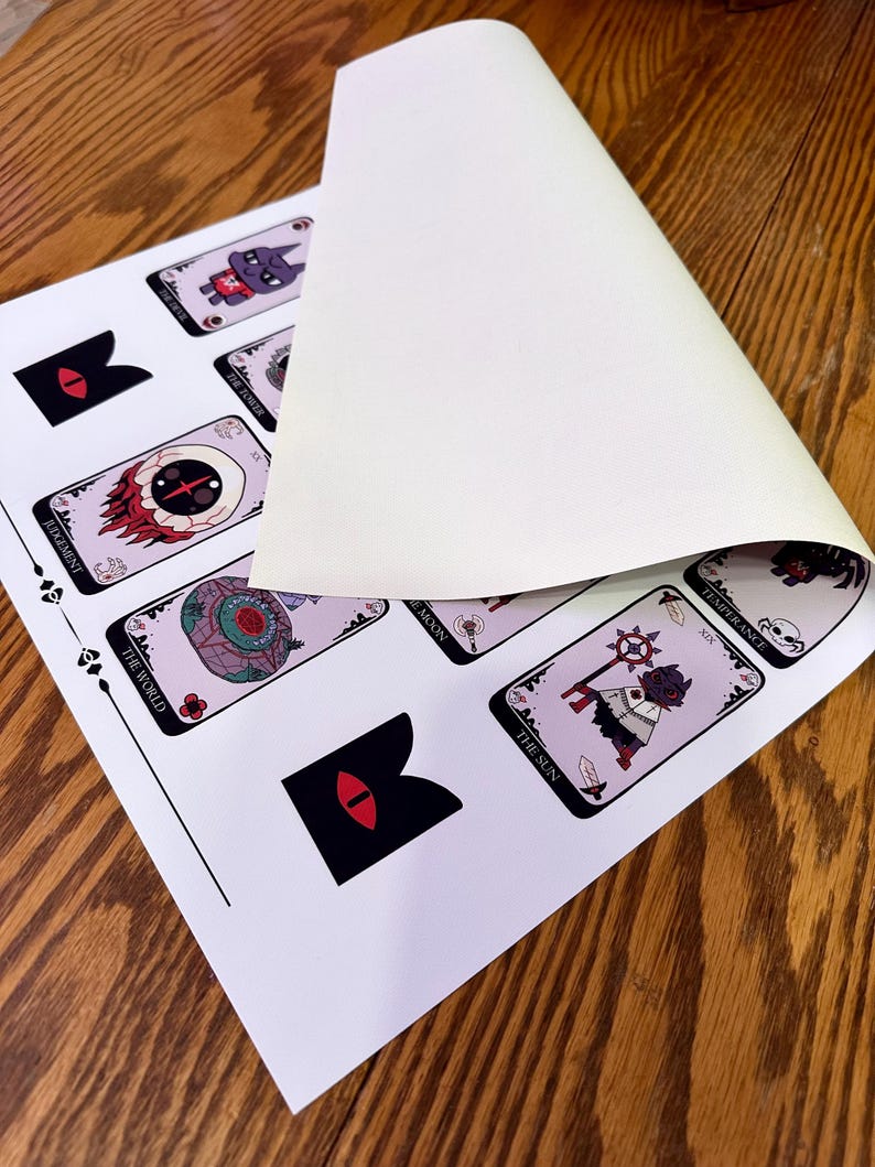 May include: A set of tarot cards featuring illustrations of various characters and symbols. The cards are printed on white paper with black borders. The cards are laid out on a wooden surface.
