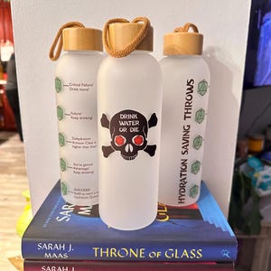 Może przedstawiać: Trzy białe, matowe szklane butelki z wodą z brązową drewnianą pokrywką i brązowym sznurkowym uchwytem. Butelki mają różne wzory. Jedna butelka ma czaszkę i skrzyżowane kości z tekstem "Drink Water or Die". Inna butelka ma tekst "Hydration Saving Throws" z zielonymi symbolami kości. Trzecia butelka ma zielony symbol kości i tekst, który brzmi: "Critical Failure! Drink more!"  "Failure! Keep drinking!"  "Dehydration Armour Class is higher than that!"  "You've gained Advantage! Keep drinking!"  "SUCCESS! Roll to start a Hydration Quest!"