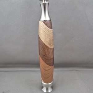 May include: A wooden pepper mill with a stainless steel top and base. The body of the mill is made of layered wood in shades of brown and tan. The mill is approximately 8 inches tall.