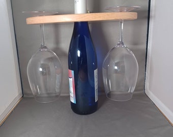May include: A wooden wine glass and bottle holder. The holder has a light wood finish and holds two wine glasses and a blue wine bottle. The wine glasses are clear and the bottle has a silver cap.